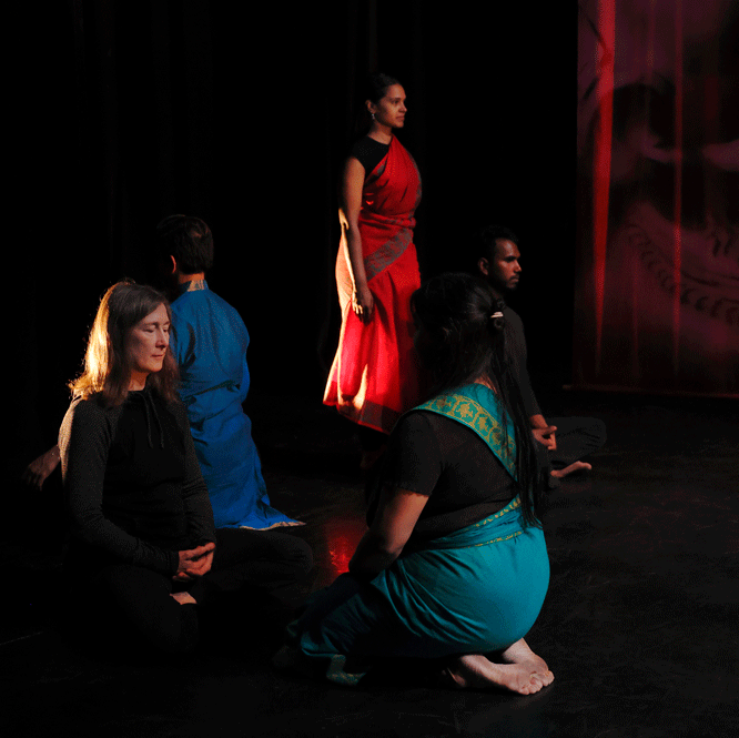 Four people kneel on a dark stage while a dance artist stands between them. They are wearing traditional Bharatanatyam clothing.