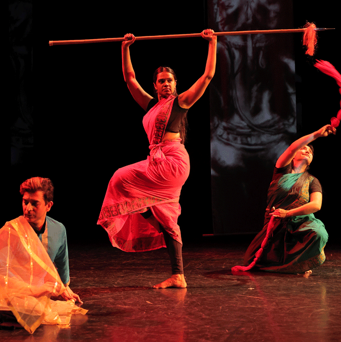 Three dance artists pose on stage. Two are low to the ground, while the middle dancer stands on one leg while holding a long stick above her head.
