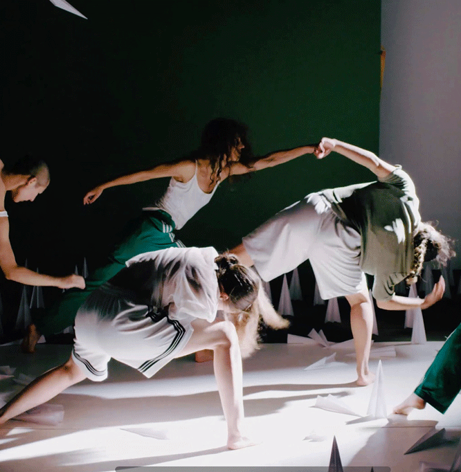 Four dancers lean forward with their arms outstretched. They are wearing white and green clothing. on the floor are numerous paper air planes standing upright or lying flat as if they had just landed.