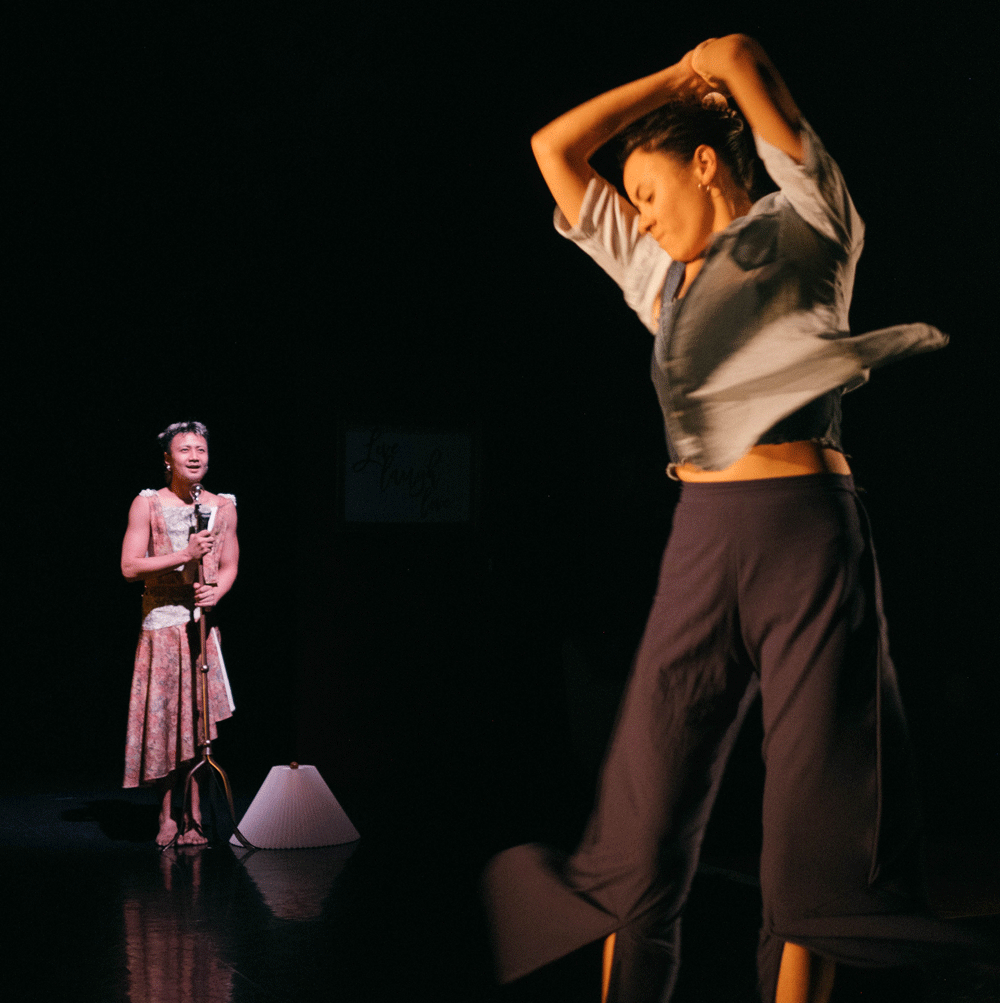 A dance artist mid twirl on a dark stage. At the back left of the image another artist stands at a mic stand wearing a pink and white dress.