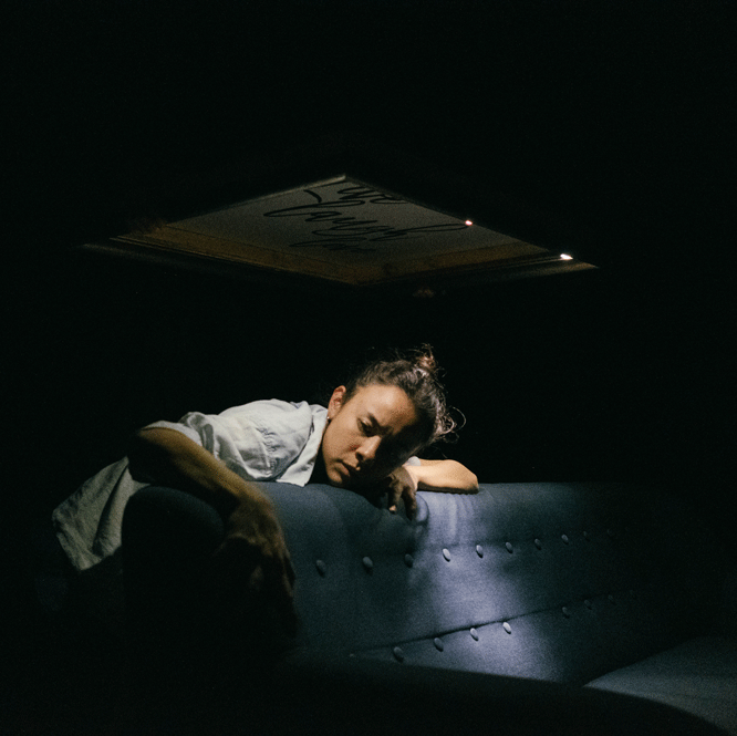 A dance artist leans over a blue couch. The image is dark except for one bright spotlight which is shining just next to the dancer lighting their face slightly.