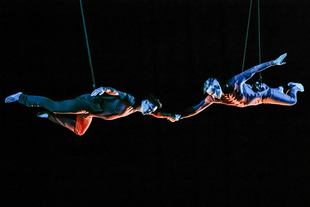 Two aerial dance artists hang horizontally with their left hands clasped.