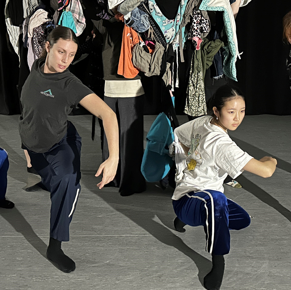 Two dance artists pose in deep lunges. They are in a dance studio with bright lights shining on their faces. Behind them are other dancers holding up many articles of clothing obscuring their faces.