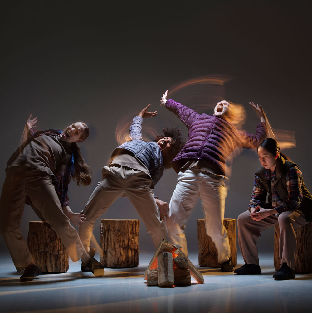 Three dance artists leaning back with their mouths open in mock screams. One other dance artist sits on a stump looking at the fake fire. They are wearing outdoor clothing and the stage is set up like a camping area.