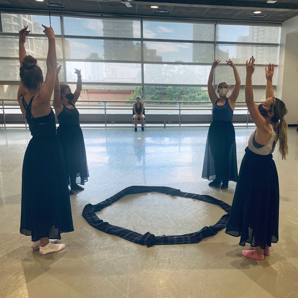 Dance artists from Shot of Scotch hold their arms above their heads while standing in a circle in a dance studio