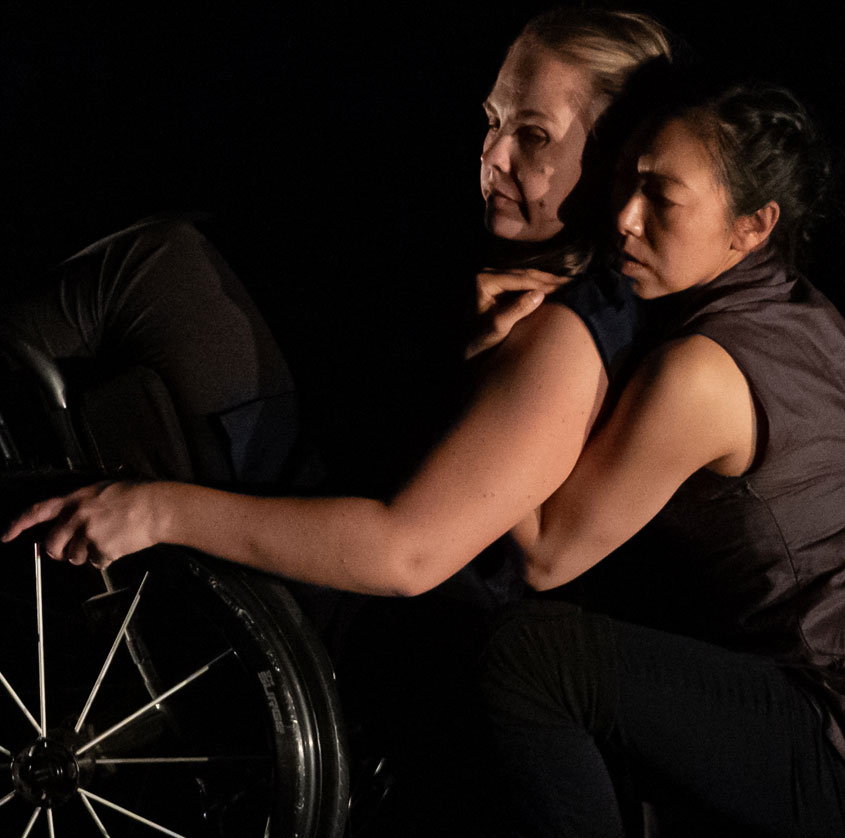 A dance artist in a wheelchair leans back and is held by another dance artist mid performance