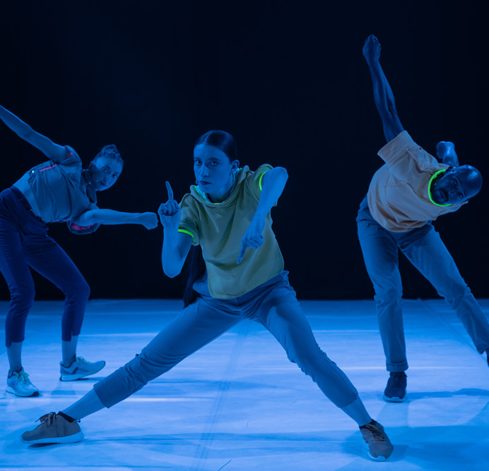 Three dance artists wearing bright clothes pose mid performance. They have glow sticks around their necks and arms and are wearing bright running shoes on the white dance floor.