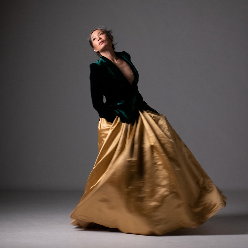 A dance artist poses. She is wearing a long gold skirt and a green blazer. Her hands are in her pockets and she is looking up and away from the camera.