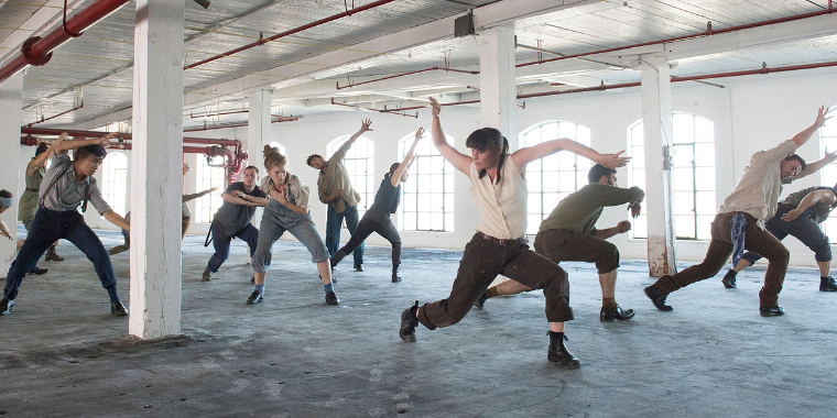 A group of dance artists in street clothing perform in a large room. the room is carpeted with large windows and exposed pipes.